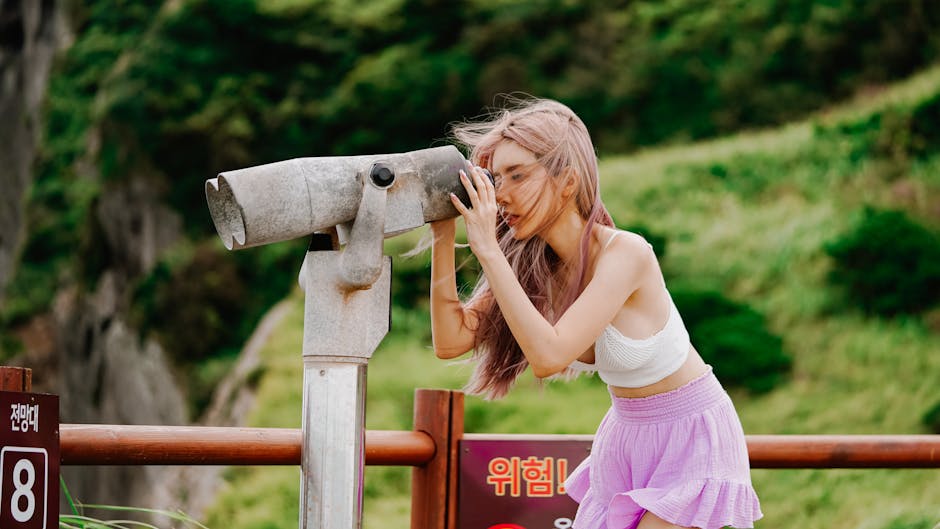 A woman with long dyed hair looks through binoculars in a scenic outdoor setting.