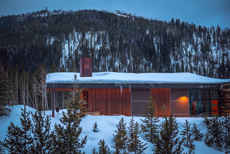 A modern cabin in a snowy forest at twilight, surrounded by mountains.