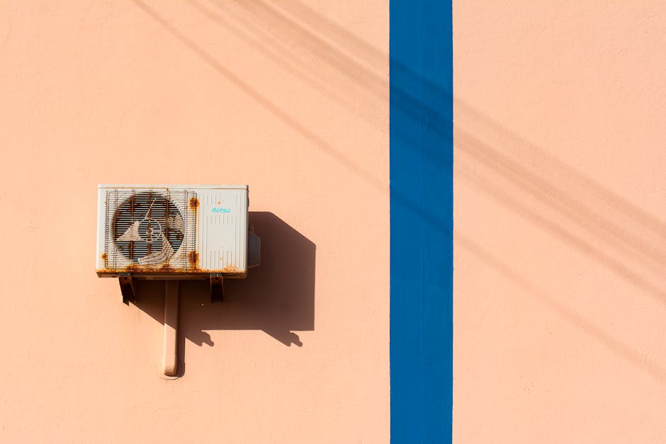 Rusty air conditioning unit casting shadow on a peach wall with blue stripe.